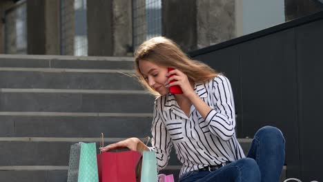 girl sitting on stairs with bags talking on mobile phone about sale in shopping mall in black friday