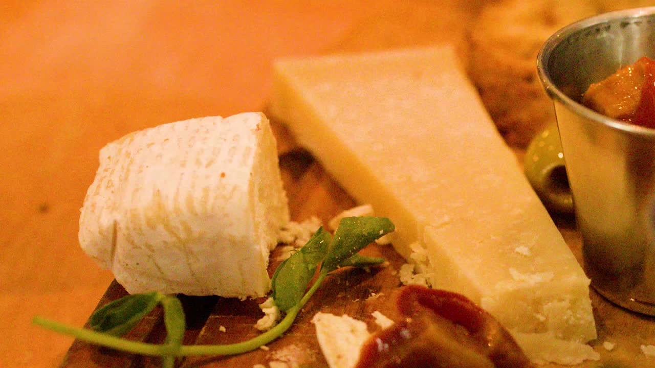 Hand slices soft cheese on rustic charcuterie board with pickles, cold cuts, and warm lighting