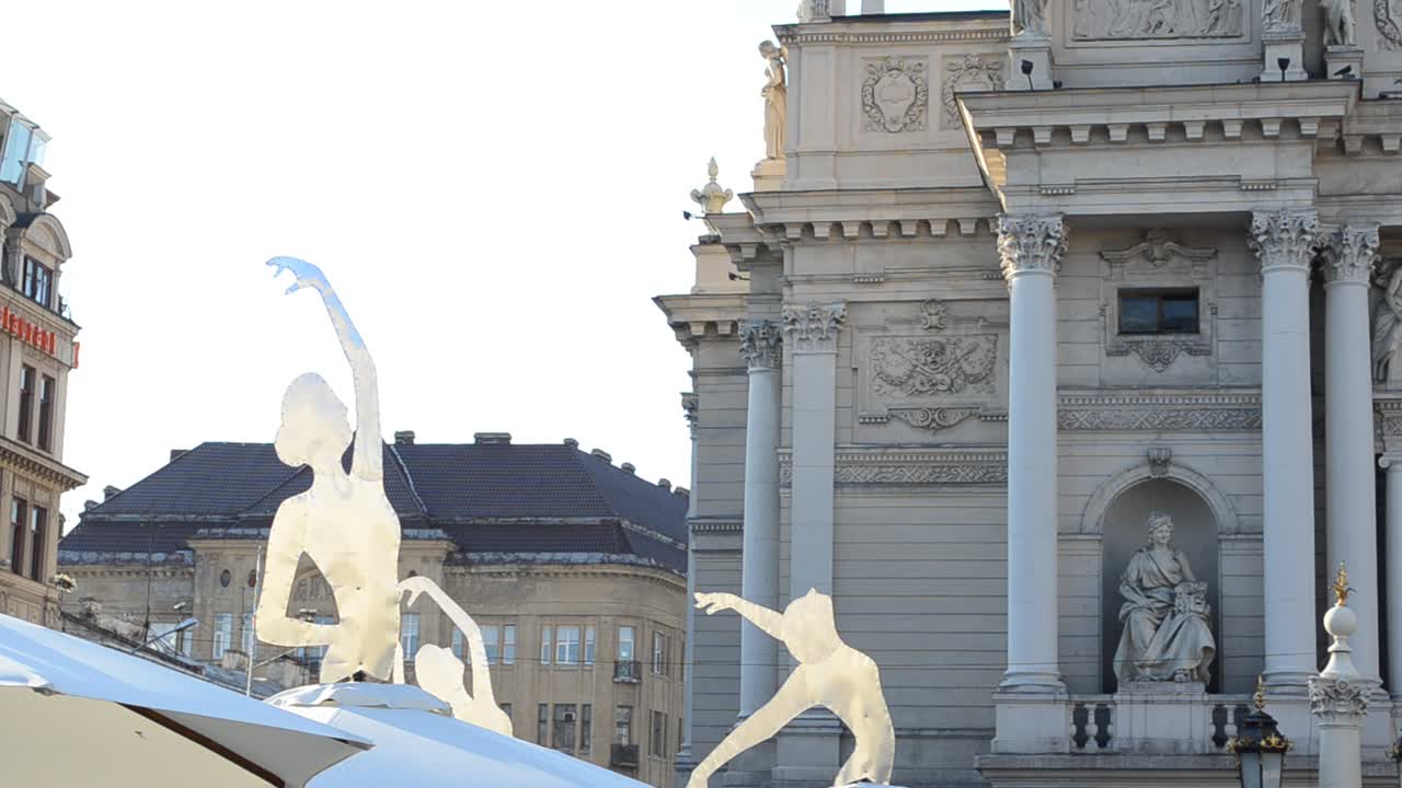 Ombrelli a forma di ballerine, caffè e la fontana. Esterno del Teatro dell'Opera e del Balletto di Leopoli. La scultura centrale è la Vittoria, quella a sinistra è la Musica, la figura a destra è la Commedia e il Dramma. Leopoli, Ucraina