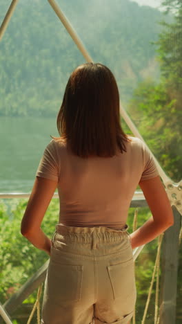 dreaming woman enjoys nature from modern window of house in forest. picturesque view of lake and mountains with dense trees. unforgettable summer holiday in clear weather