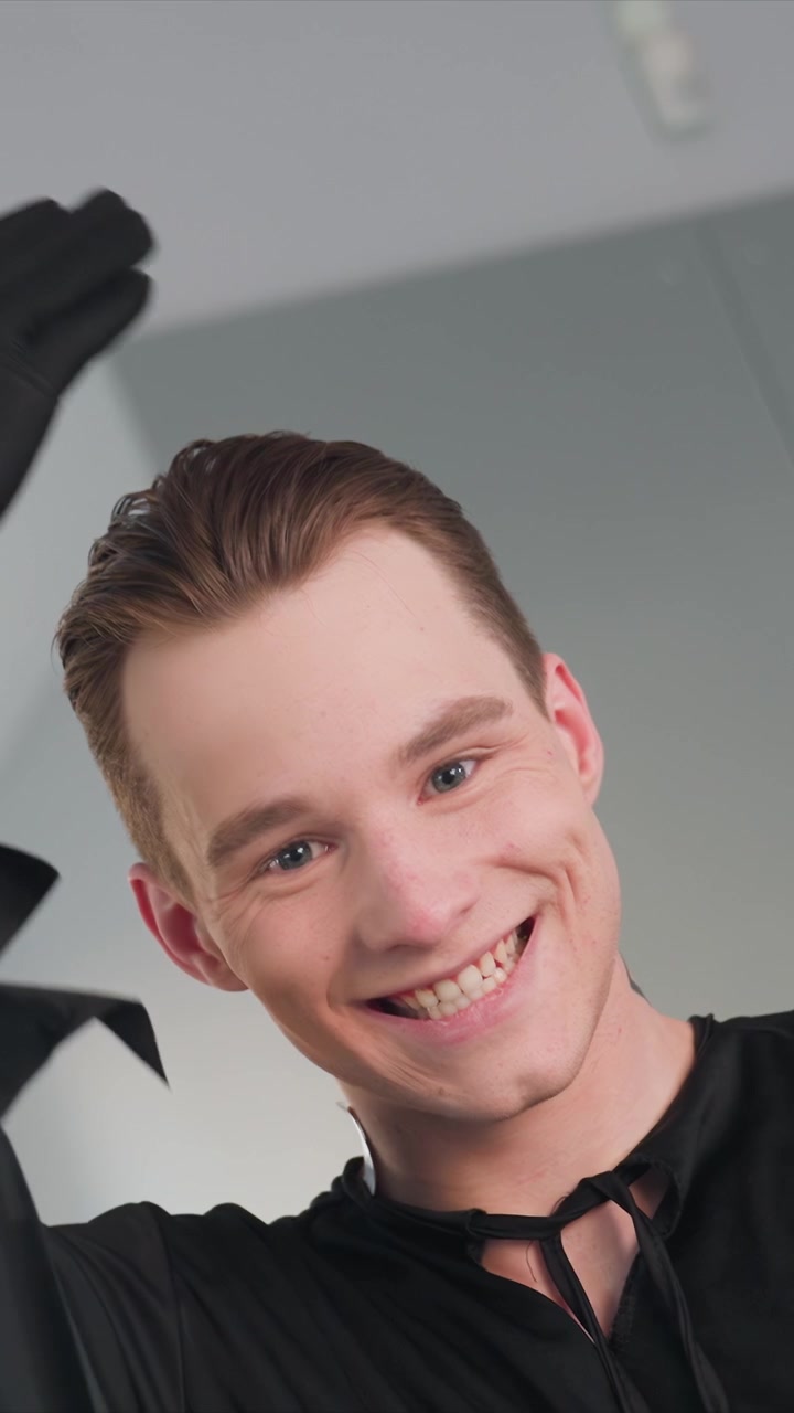 Young man with brown hair styles his neat haircut using gloved hands. Man wears black top featuring tied strings. Background is light colored walls and ceiling suggesting interior
