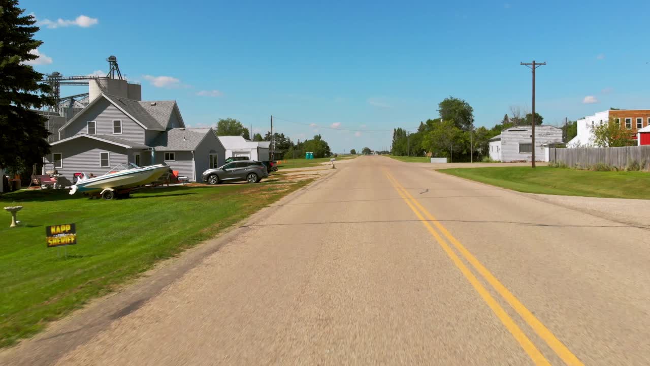 Low aerial shot flying backward along a quiet small-town street lined with houses, trees, and power poles under a clear blue sky