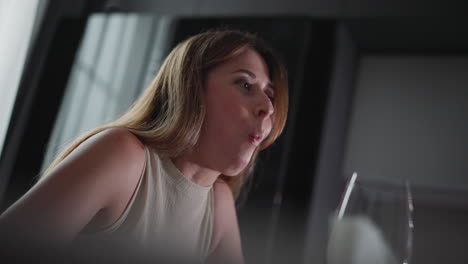 young lady with long hair slightly bent forward, eagerly loading her mouth with food using chopsticks while sitting at dining table, wearing sleeveless top