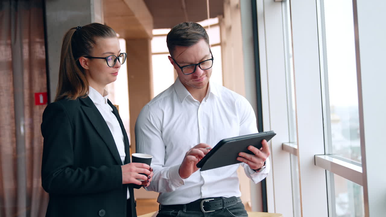 Caucasian coworkers standing near the window look at the I-pad in male hands. Woman with coffee cup in hand talks to man and points on gadget.