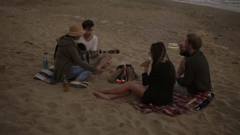 young friends sitting by the fire on the beach, grilling sausages, drinking beer and playing guitar. slow motion shot