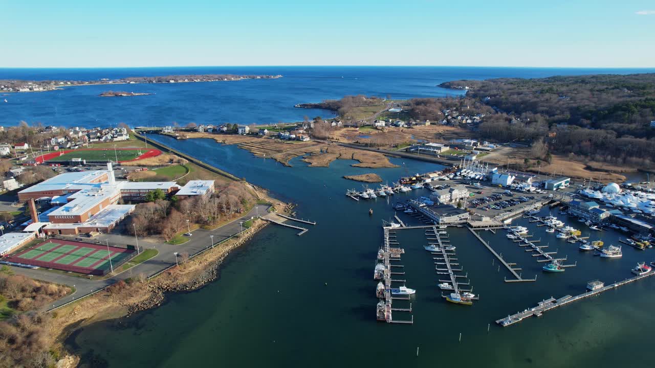 Vista aerea sopra il porto turistico e il porto di Gloucester, Massachusetts, Stati Uniti. I moli della marina e le barche fiancheggiano le calme vie d'acqua blu accanto alla città costiera sotto cieli limpidi e luminosi. Orbit