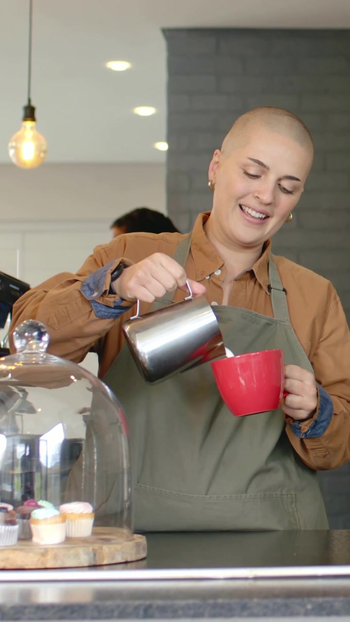 Video verticale: Un barista con grembiule color oliva che tiene una brocca d'acciaio e una tazza rossa, versando il latte al bancone, con cupcake.