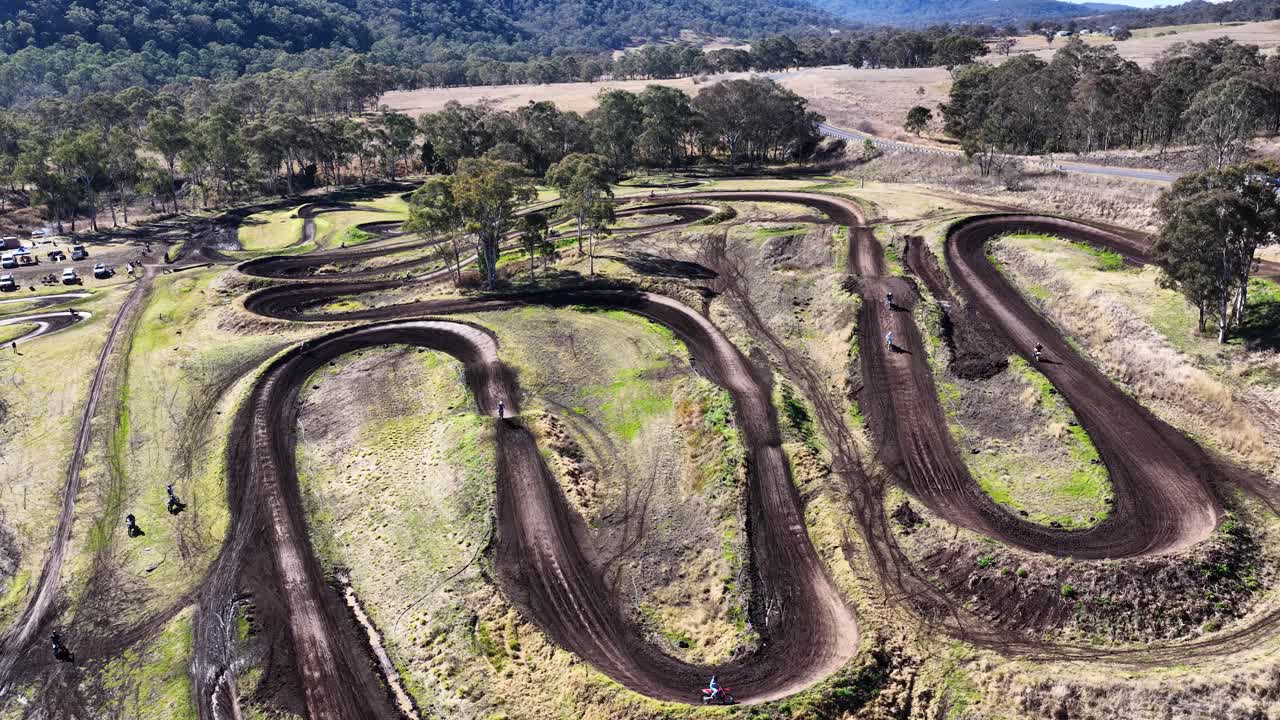 Drone footage tracks motocross riders navigating winding dirt course under bright daylight in rural Australia