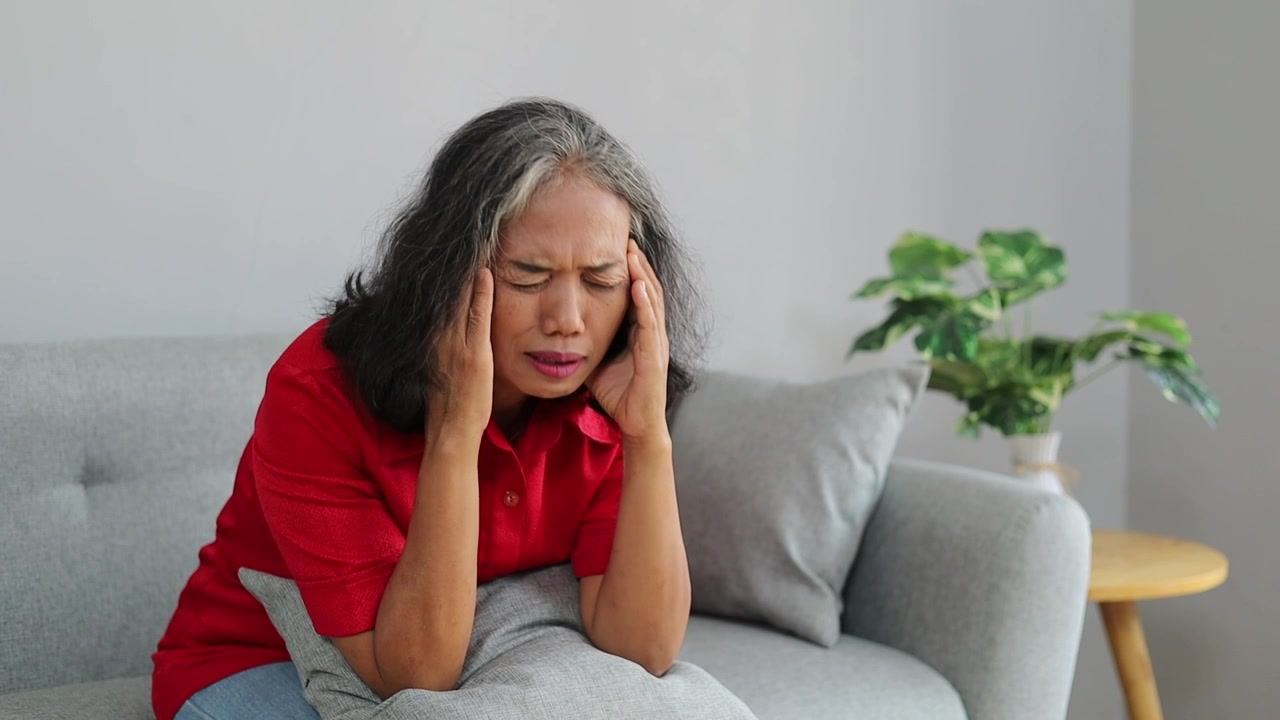 Asian senior woman sitting on a sofa, holding her temples with eyes closed in distress, illustrating headache, migraine, dizziness, stress, fatigue, and health problems.