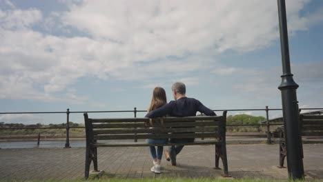 dad and daughter sit together on bench chatting and cuddling, wide shot