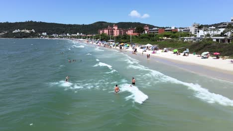 escena aérea de drones volando bajo sobre el mar y olas con mucha gente buceando y divirtiéndose en la arena de la playa con muchos edificios y hoteles frente al mar en florianópolis