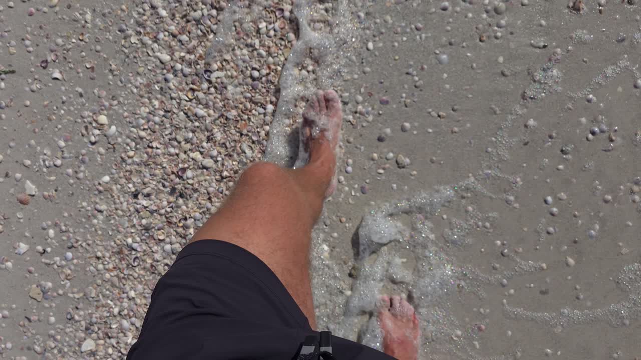 A barefoot person strolls along a sandy beach scattered with seashells and small bubbles of sea foam. The scene highlights a relaxed coastal moment.