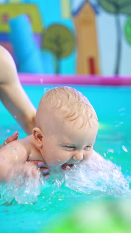 adorable toddler splashing water actively in the swimming pool. baby is taught to swim by a female trainer. vertical video.
