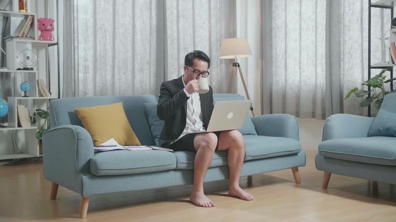 집에서 노트북으로 일하면서 커피를 마시는 재과 쇼트를 입은 아시아 사업가