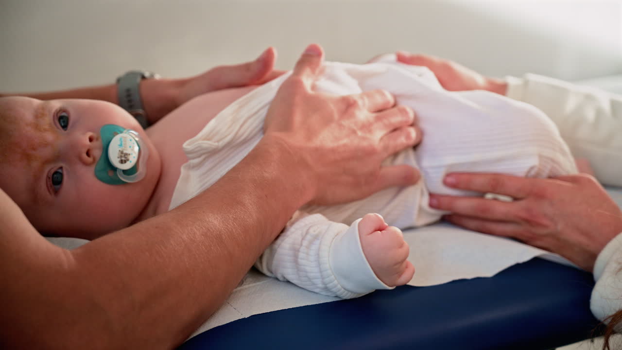 Primo piano di un neonato disteso tranquillamente su un lettino medico, che tiene in mano un ciuccio mentre un adulto lo sostiene delicatamente