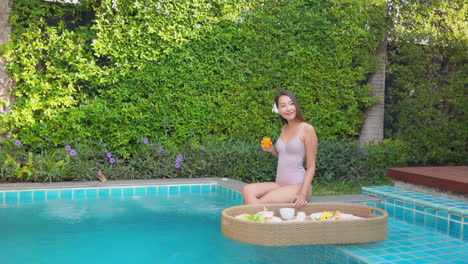 slow-motion on slim asian woman holding a glass of orange juice and ready to eat breakfast from floating on the water tray poolside