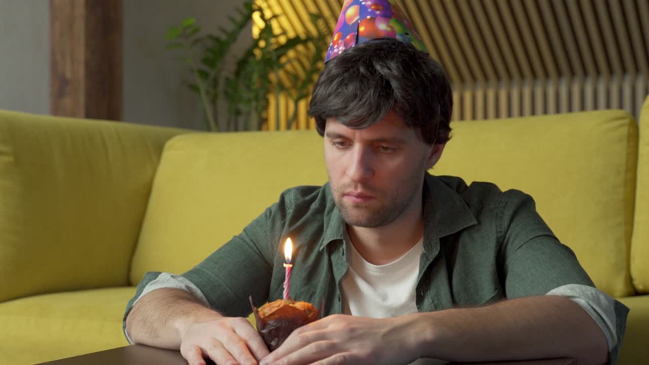 hombre triste celebra su cumpleaños solo en la sala de estar, soplando las velas en el pastel