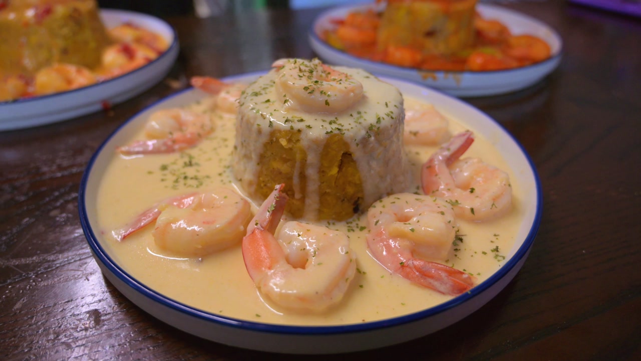 Cinematic close up shot of a traditional Puerto Rican mofongo. Mashed green plantains topped with shrimp in a creamy garlic sauce served in a wooden bowl at a restaurant.
