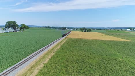 an aerial view of a steam engine puffing smoke and steam with passenger coaches traveling on a single track fertile farmland and countryside on a beautiful spring day