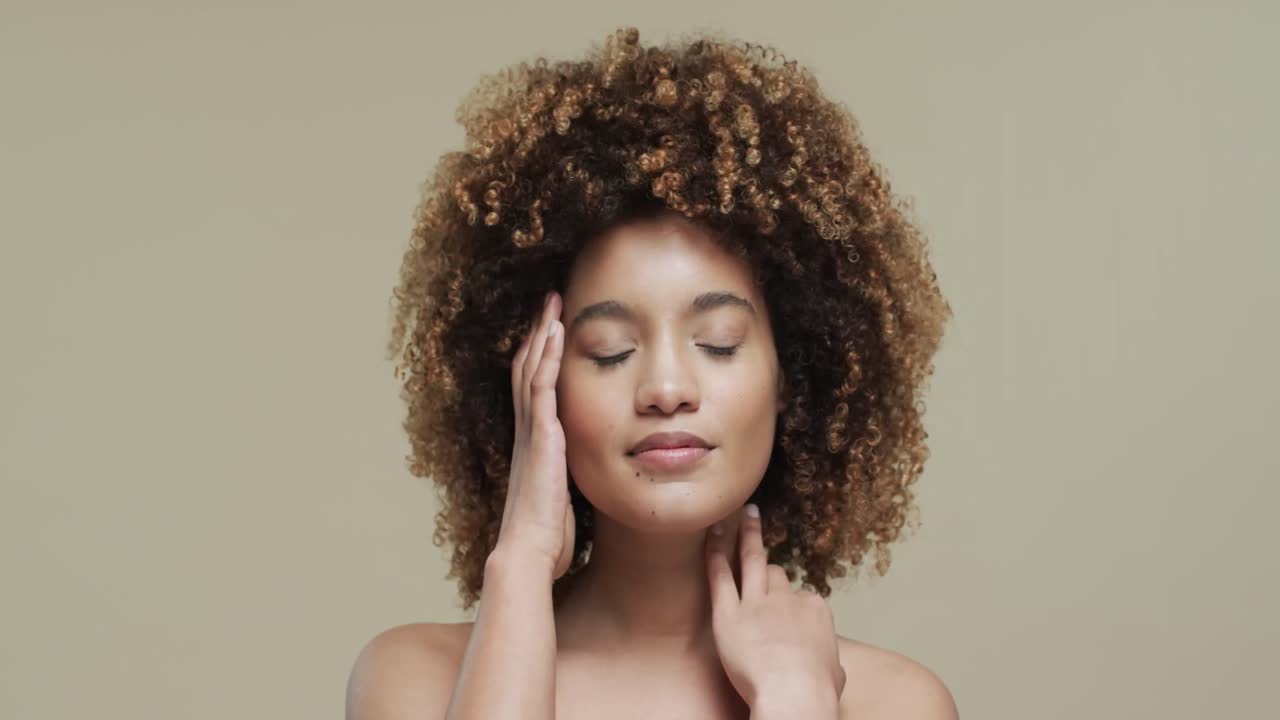 Biracial woman with dark hair touching face on beige background with copy space, slow motion