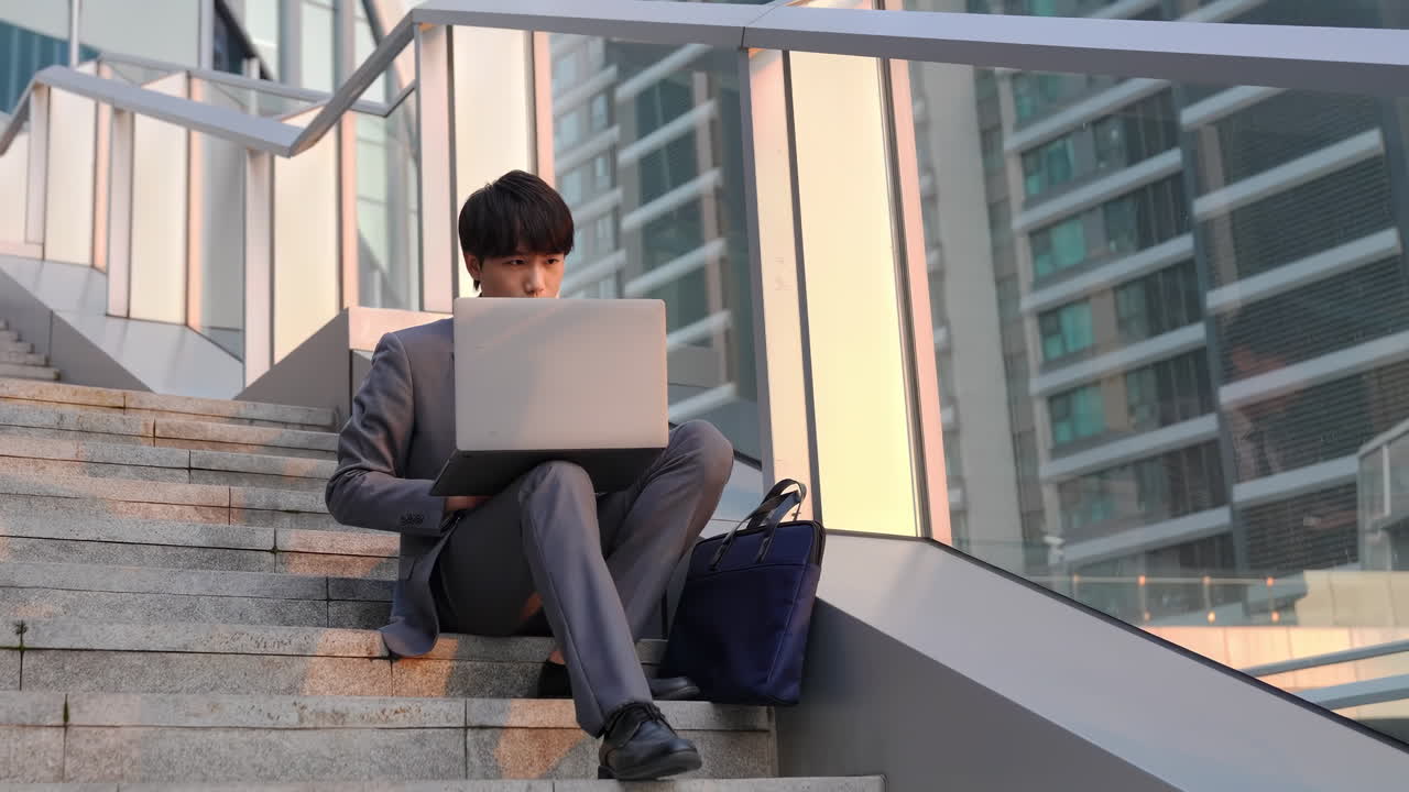 Drukke zakenman in pak werkt met laptop terwijl hij op een trap zit bij een kantoorgebouw, naast een aktetas. Freelancer werkt aan een project in een stedelijke buitenruimte, gebruikt de computer en controleert de toenemende verkoop.