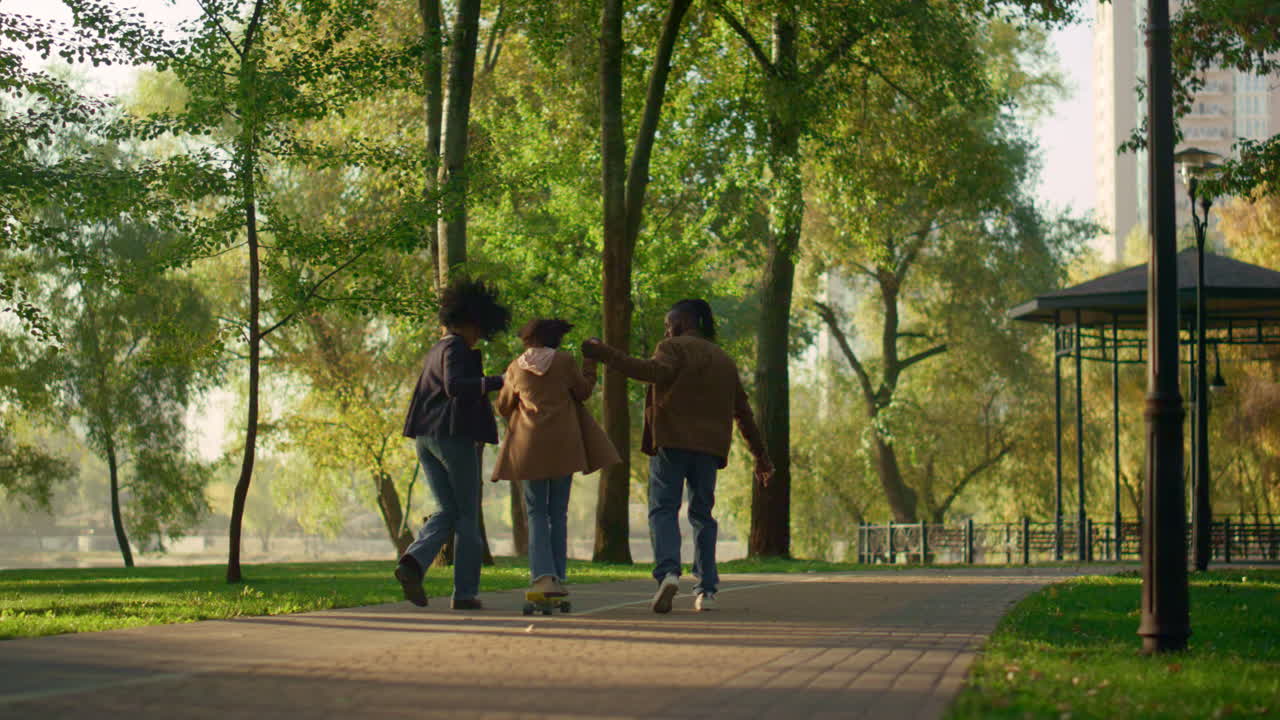 롱보드를 타고 있는 소녀가  ⁇ 은 가을 공원의 뒷면에서 부모의 손을 잡고 있습니다.