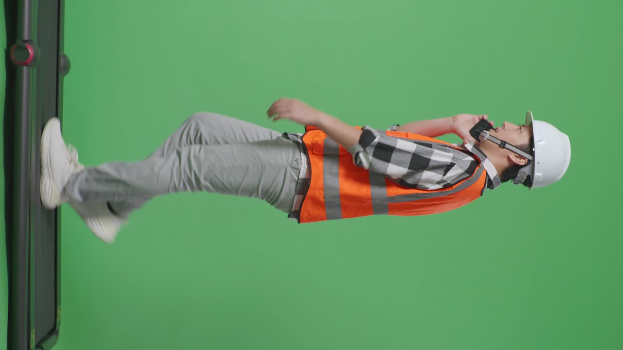 vista lateral de cuerpo completo ingeniero masculino asiático con casco de seguridad hablando en teléfono inteligente mientras camina en el estudio de fondo de pantalla verde