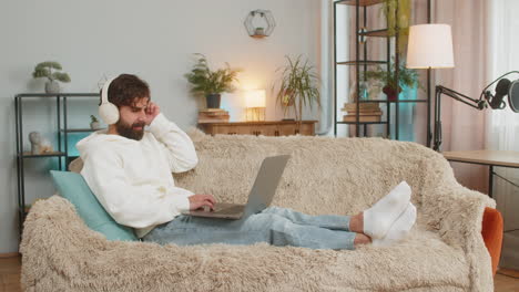 happy relaxed indian young man in wireless headphones listening to music using laptop on sofa couch