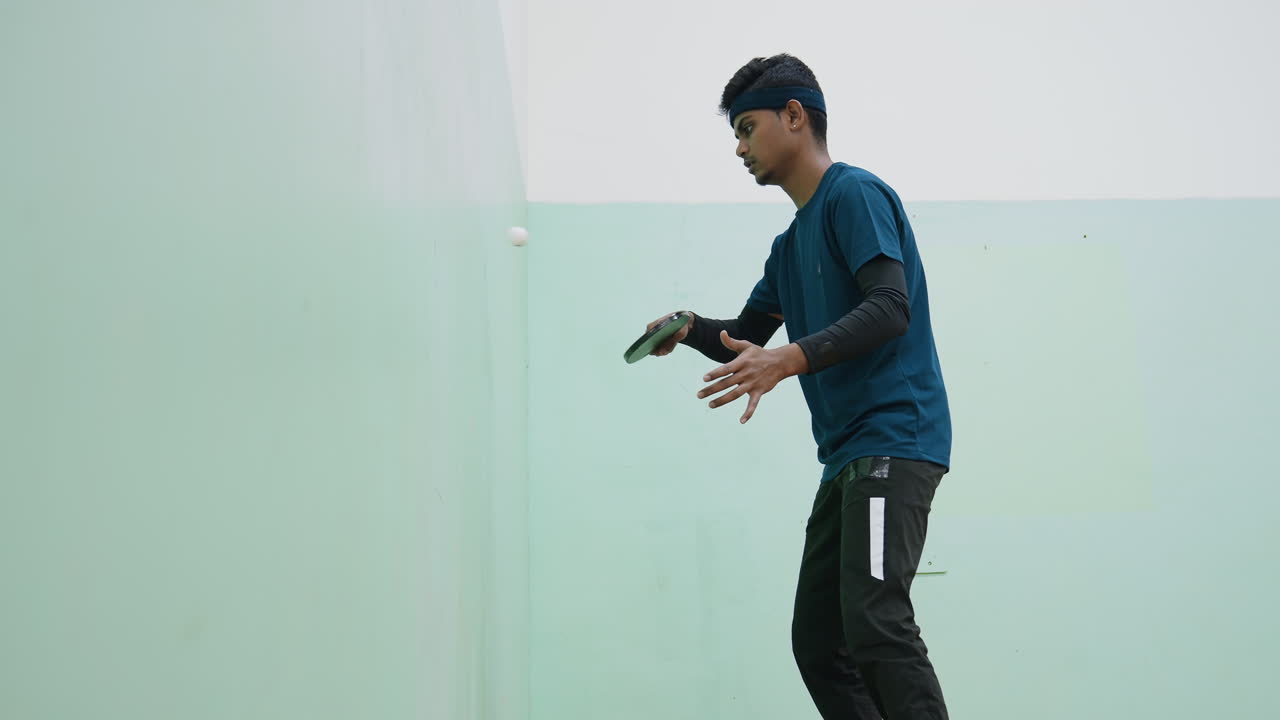 Tenis oyuncusunun içeride duvara karşı antrenman yaparken yandan görünümü, raketi odaklanmış bir şekilde tutarak, topa kontrollü hareketlerle vururken, spor kıyafetleri ve saç bandı takıyor.