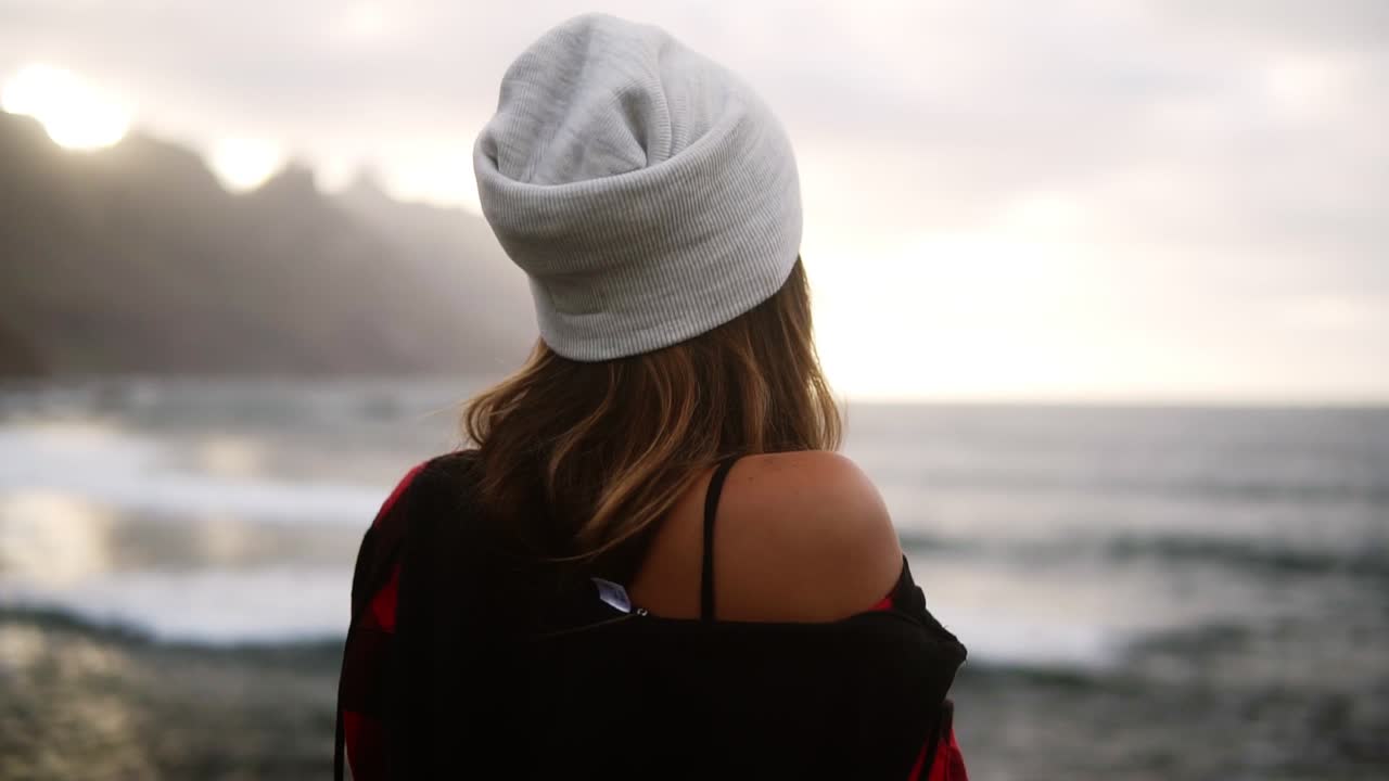 Woman gazes at ocean - A brunette woman shown from behind looks out over an ocean in front of the hills, and then turns her head to the side smiling over her shoulder. Blurred background