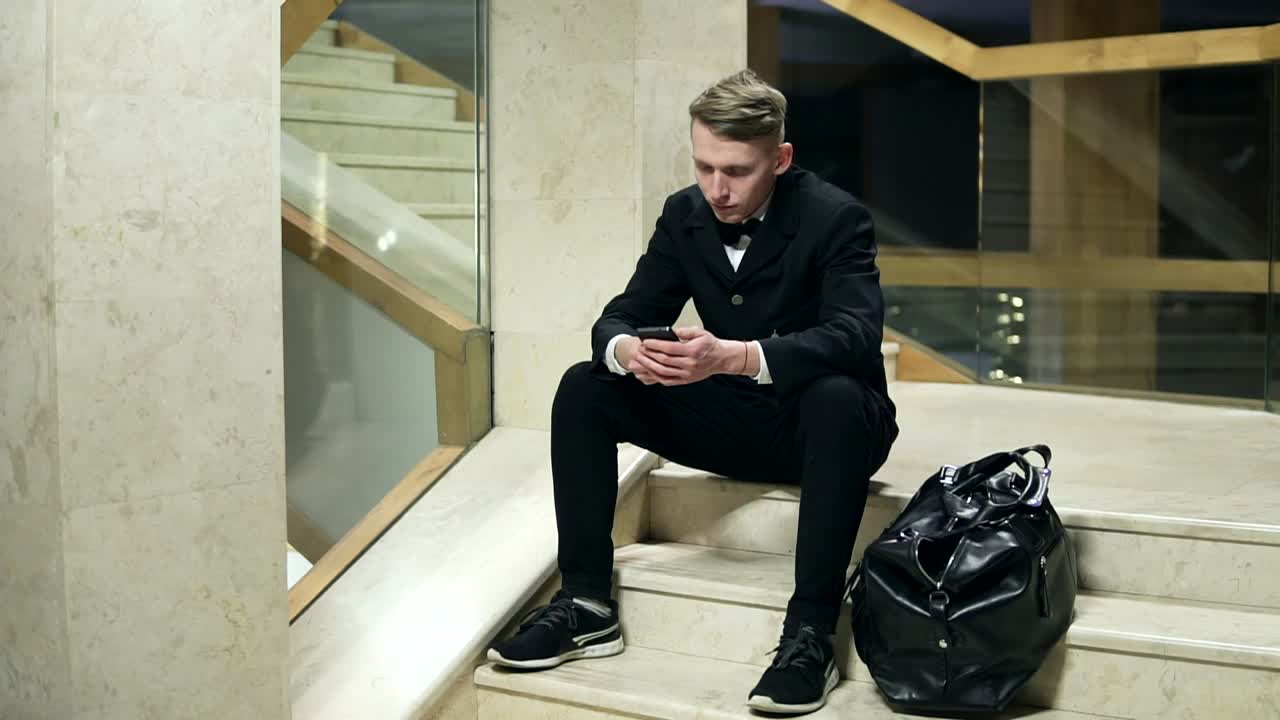 Young blonde man in black suit with big black leather bag using his smartphone and sitting on stairs in hotel. Dialing a number