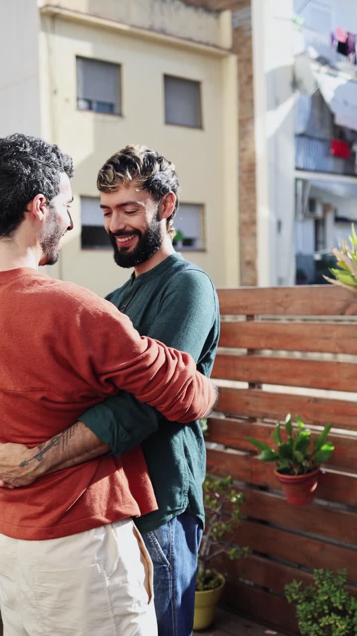 Coppia gay felice che si abbraccia e sorride su una terrazza soleggiata.