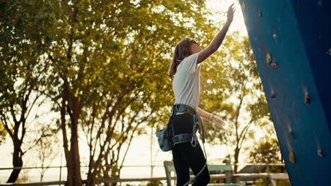 una chica rubia escaladora en una camiseta blanca saca tiza deportiva, la frota y comienza a escalar la pared de escalada azul en un soleado día de verano