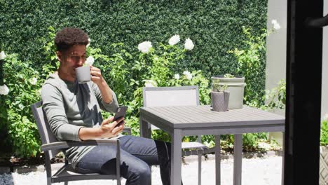 hombre biracial feliz usando un teléfono inteligente y bebiendo café en el jardín