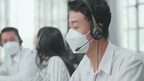Close Up Of A Man Of Three Asian Call Center Agents Wearing Headset And Mask Speaking To Customer On The Call While Two Of His Colleagues Are Talking During Working At The Office