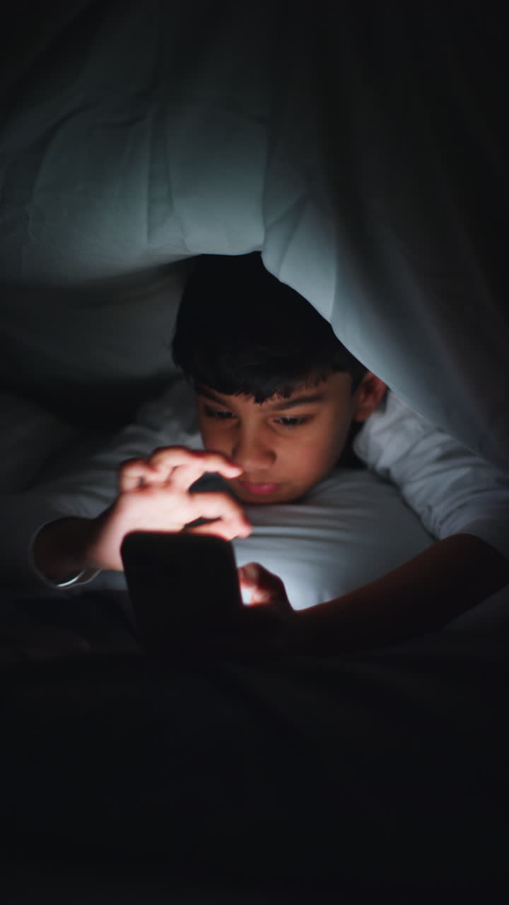 Vertical Video Close Up Of Young Boy In Bedroom At Home Using Mobile Phone To Text Message Under Covers Or Duvet At Night 1