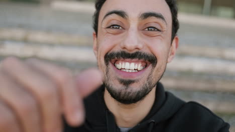 medium shot of young arabic mans face with dark curly hair and beard in black hoodie sitting on stairs outside