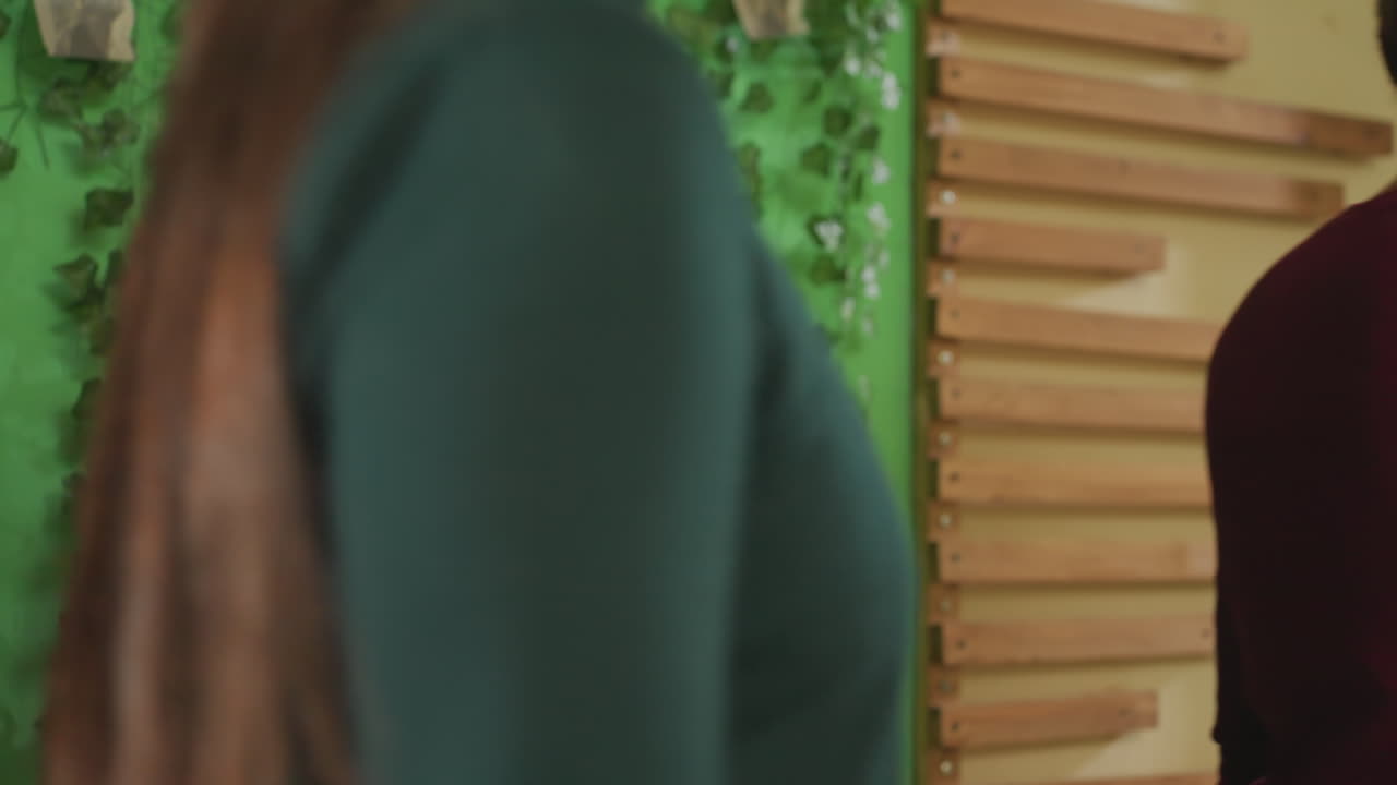 Redhaired white woman presenting green studio instructor introduces activity while young boy observes, warm interaction, wooden slat backdrop, vine foliage, casual outfit, confident stance