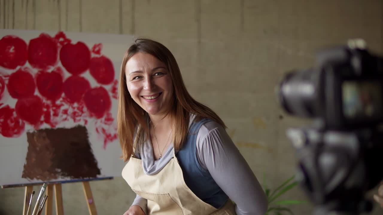 Female caucasian mature blogger recording video in workshop - drawing pink flower on canvas, explaining details on camera. Slow motion. Tripod camera on foreground