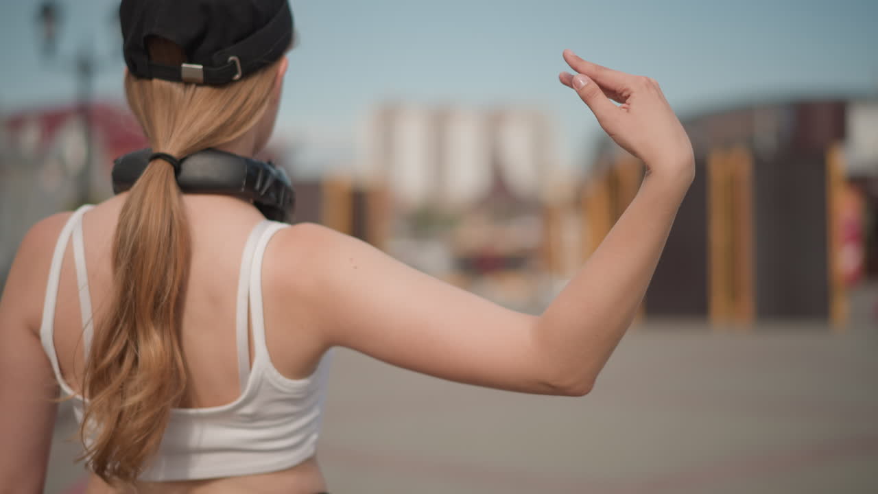 Ballerina di strada, donna bianca, vista da dietro, in movimento, con cuffie e berretto, coda di cavallo che oscilla mentre prova la coreografia su una strada urbana vicino a container navali, skyline della città sfocato, top corto casual