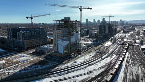 drone flying close to multi storey building under construction site with view of rail track covered with snow in and downtown denver city view in background, colorado