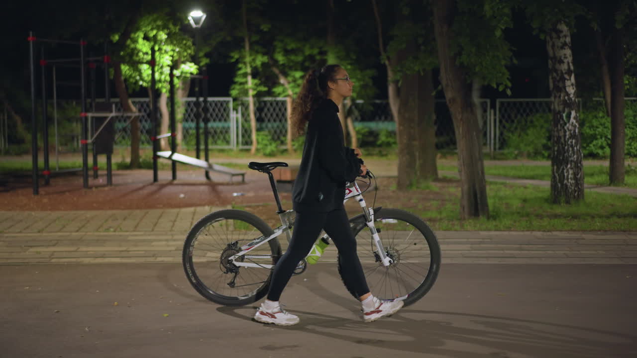Woman Cycling Through Lit Park, Caucasian Lady Navigating Bicycle Through Outdoor Illuminated Playground, Night Scene Showing Woman Riding Bicycle Under Gentle Lights Past Trees And Playground