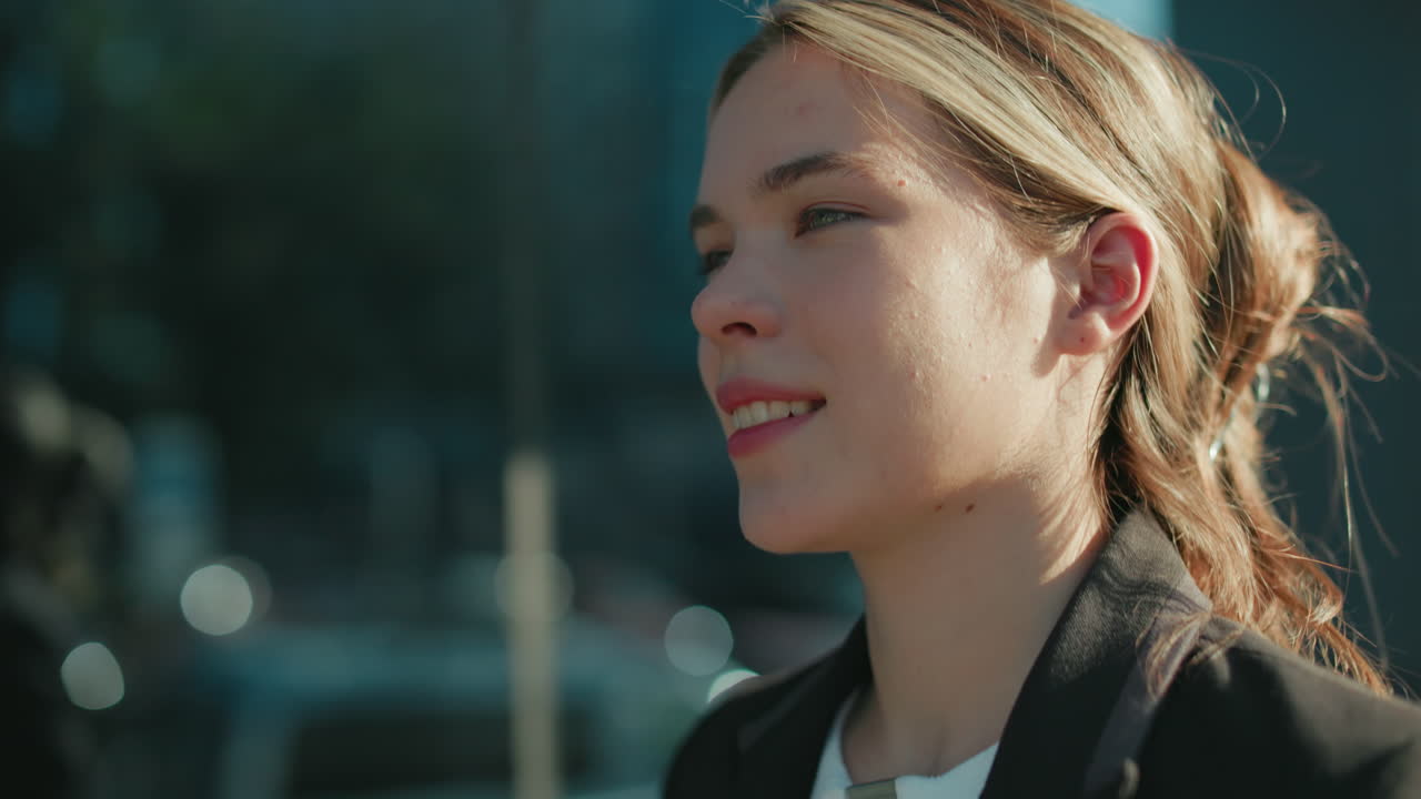Signora con un sorriso caloroso e i capelli raccolti che cammina con sicurezza accanto a una moderna struttura in vetro che riflette l'ambiente urbano circostante, con la luce del sole che proietta un bagliore delicato sul suo viso in un elegante abito professionale