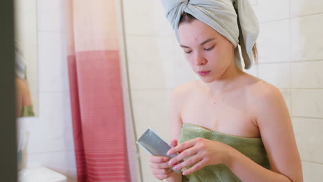 focused woman wrapped in light green towel, applying cream on her hand after shower, she is in bathroom, taking care of her skin with cosmetic products for personal hygiene and grooming