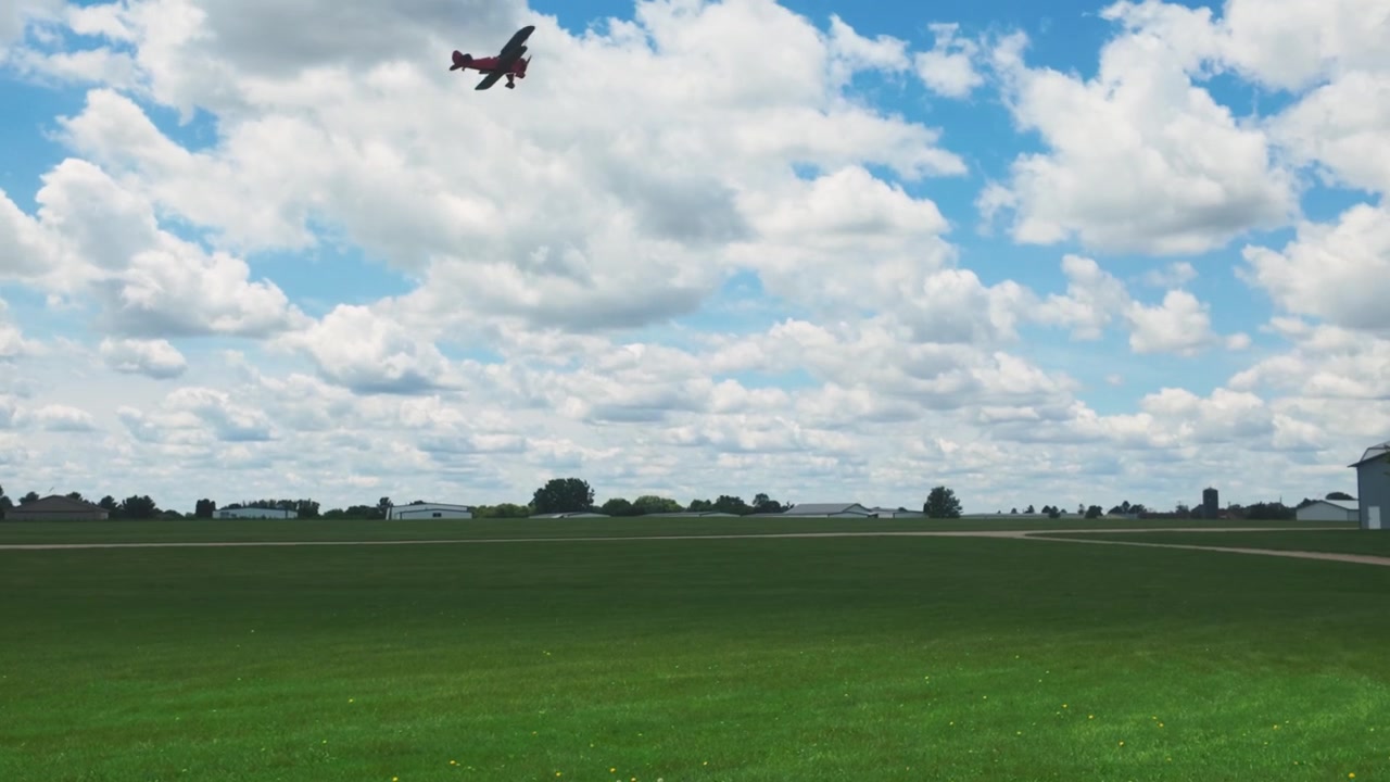 Et vintage rødt propelplan, Red Waco, der letter fra en græsmark og flyver ind i en lys himmel fuld af skyer.