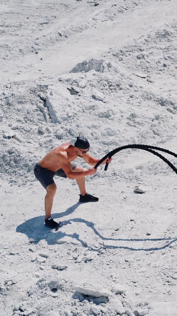 Strong young man doing exercises with a battle rope in sunny day. View from above on a muscular man training with cable on white ground outside. Concept of professional training. Vertical video