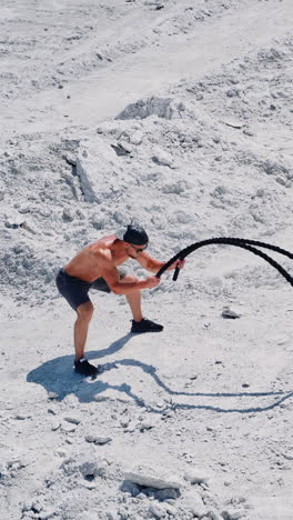 strong young man doing exercises with a battle rope in sunny day. view from above on a muscular man training with cable on white ground outside. concept of professional training. vertical video