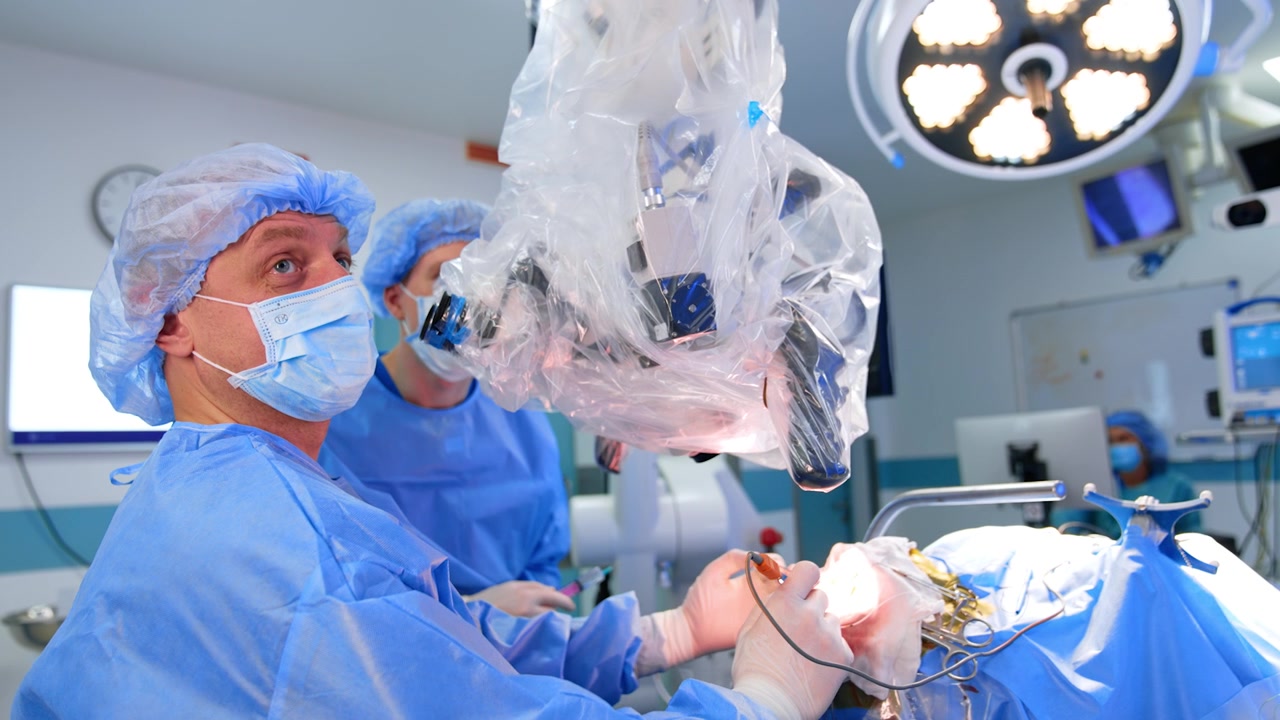 Cirurgião caucasiano com touca e máscara olha para o microscópio e aplica instrumento elétrico. Neurocirurgião trabalha na operação. Vista lateral.