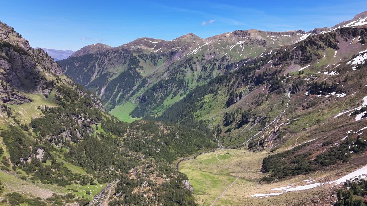 Wide mountain valley at Murgsee, Switzerland with green meadows, forested slopes, rocky ridges, and patches of snow under a clear blue sky, highlighting natural alpine scenery and rugged terrain