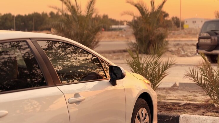 Profilo laterale in primo piano di un'auto bianca parcheggiata all'aperto durante un caldo tramonto nell'ora d'oro. I finestrini riflettono le palme circostanti, con un SUV scuro sfocato nel parcheggio polveroso sullo sfondo